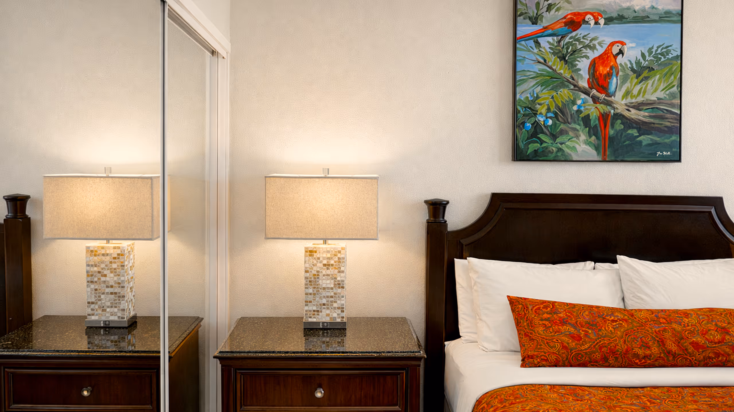 Bedroom corner with a wooden nightstand holding a mosaic-patterned table lamp and part of a dark wooden bed with white pillows and orange patterned bedding, under a painting of two red parrots on a tree branch.