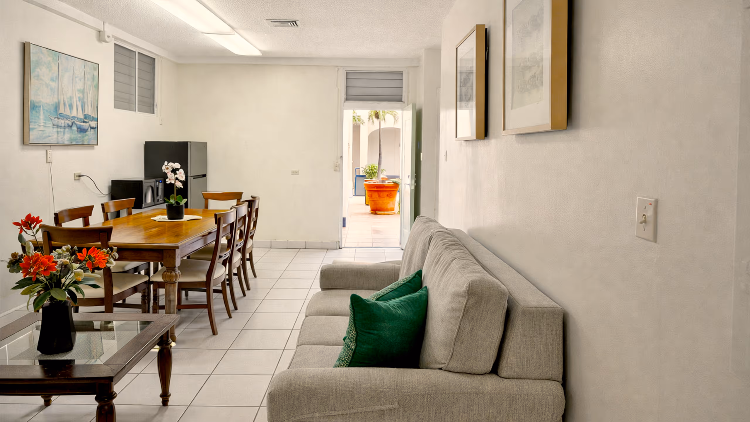 Living and dining area with beige couch with green pillows, wooden dining table with six chairs, and a glass coffee table with red flowers, leading to an open door with outdoor plants in view.