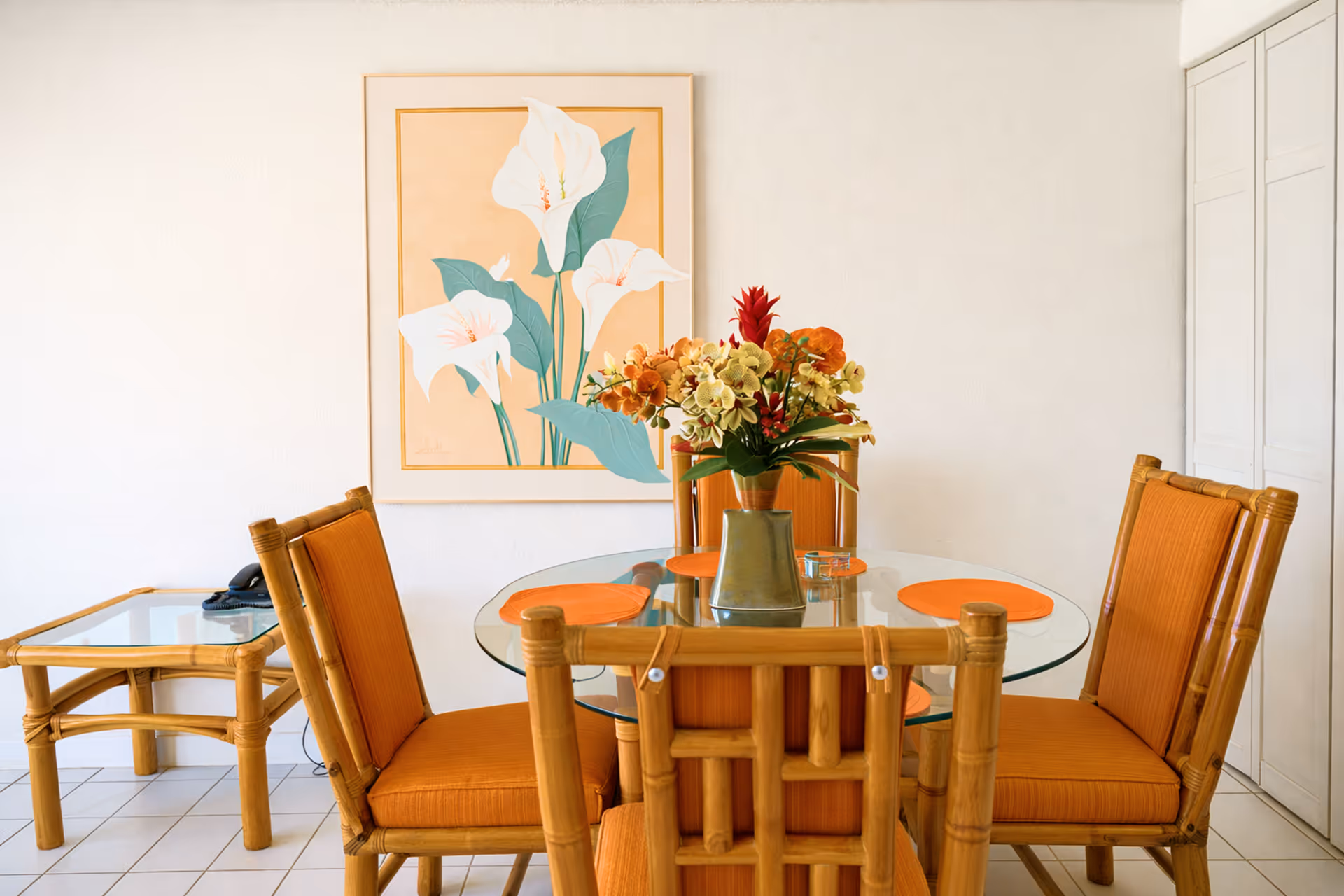 Dining area with a round glass table, four orange cushioned bamboo chairs, a flower vase centerpiece, and a wall painting of white flowers.