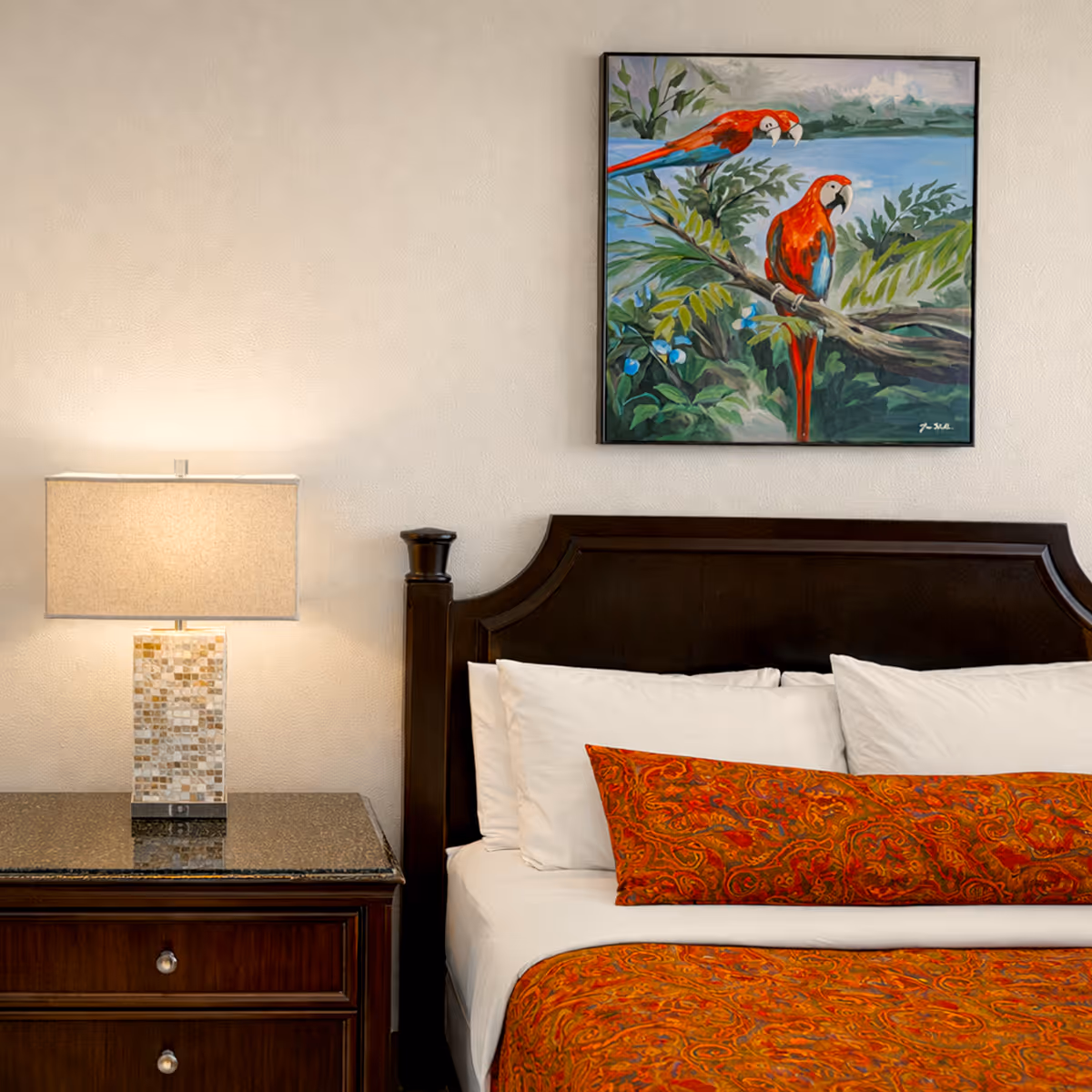 Bedroom with a dark wooden headboard bed dressed in white sheets and orange patterned bedding, next to a nightstand with a mosaic lamp and a painting of two colorful parrots on the wall above.
