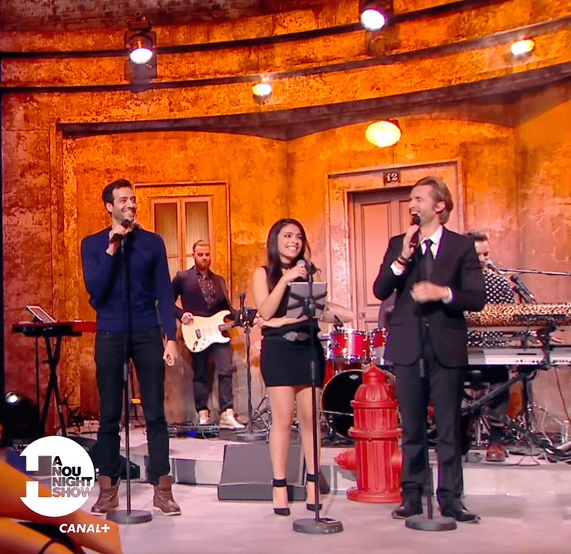 Three singers on a stage during a live music show with band members and musical instruments in the background.