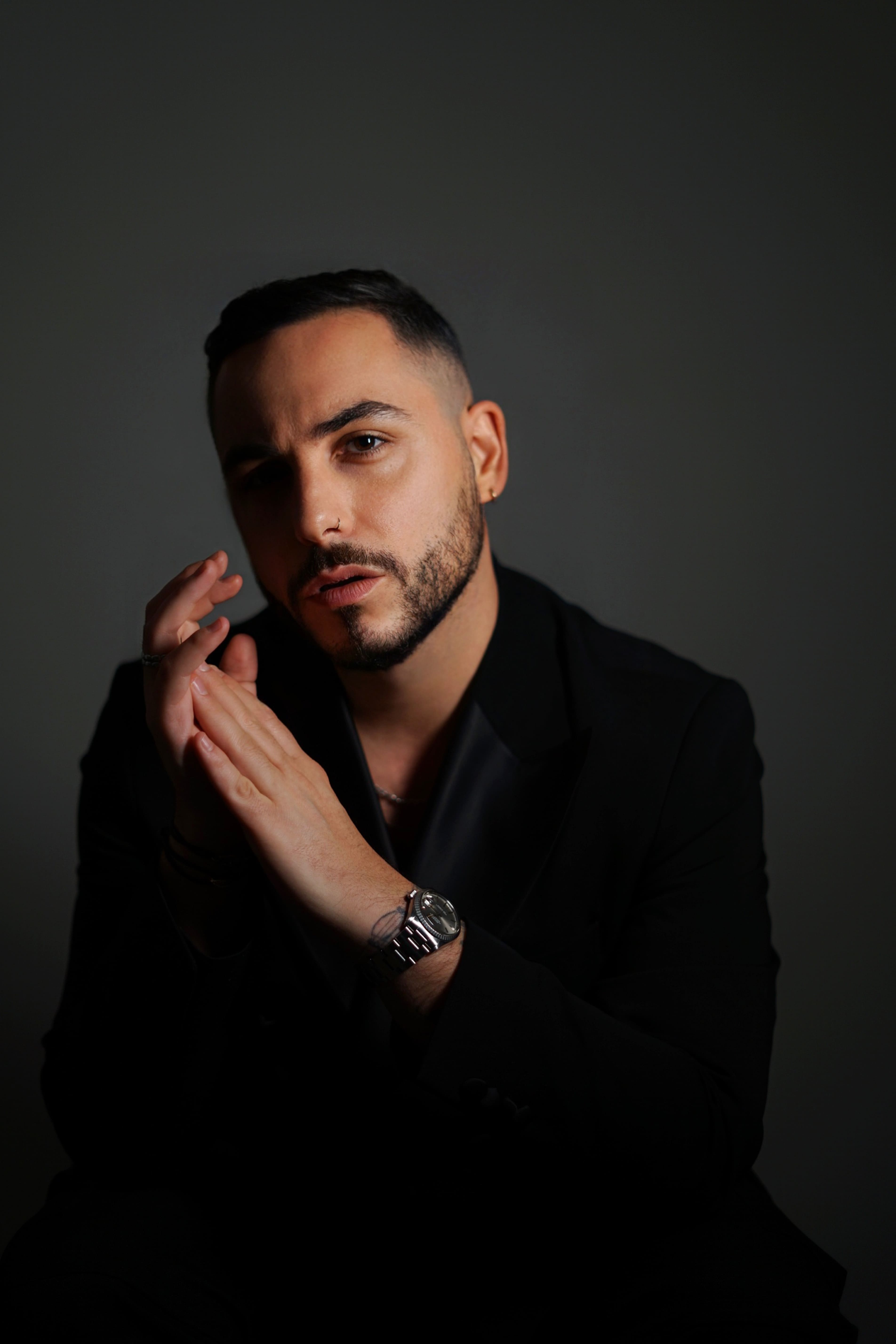 Portrait of a man with a trimmed beard and short hair wearing a black blazer and watch, posed against a dark background.