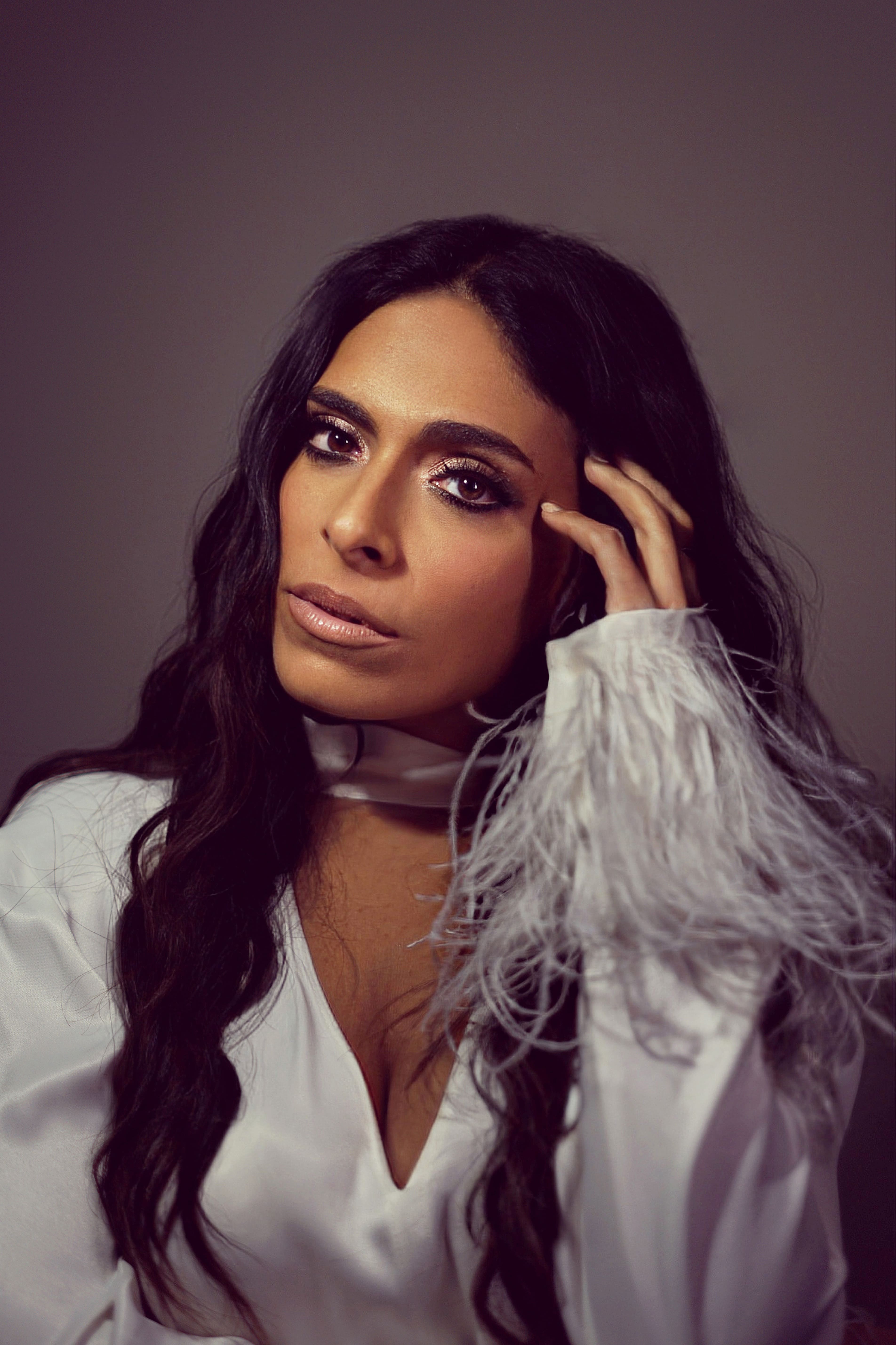 Woman with long dark hair wearing a white blouse with feathered sleeves, touching her temple and looking into the camera.