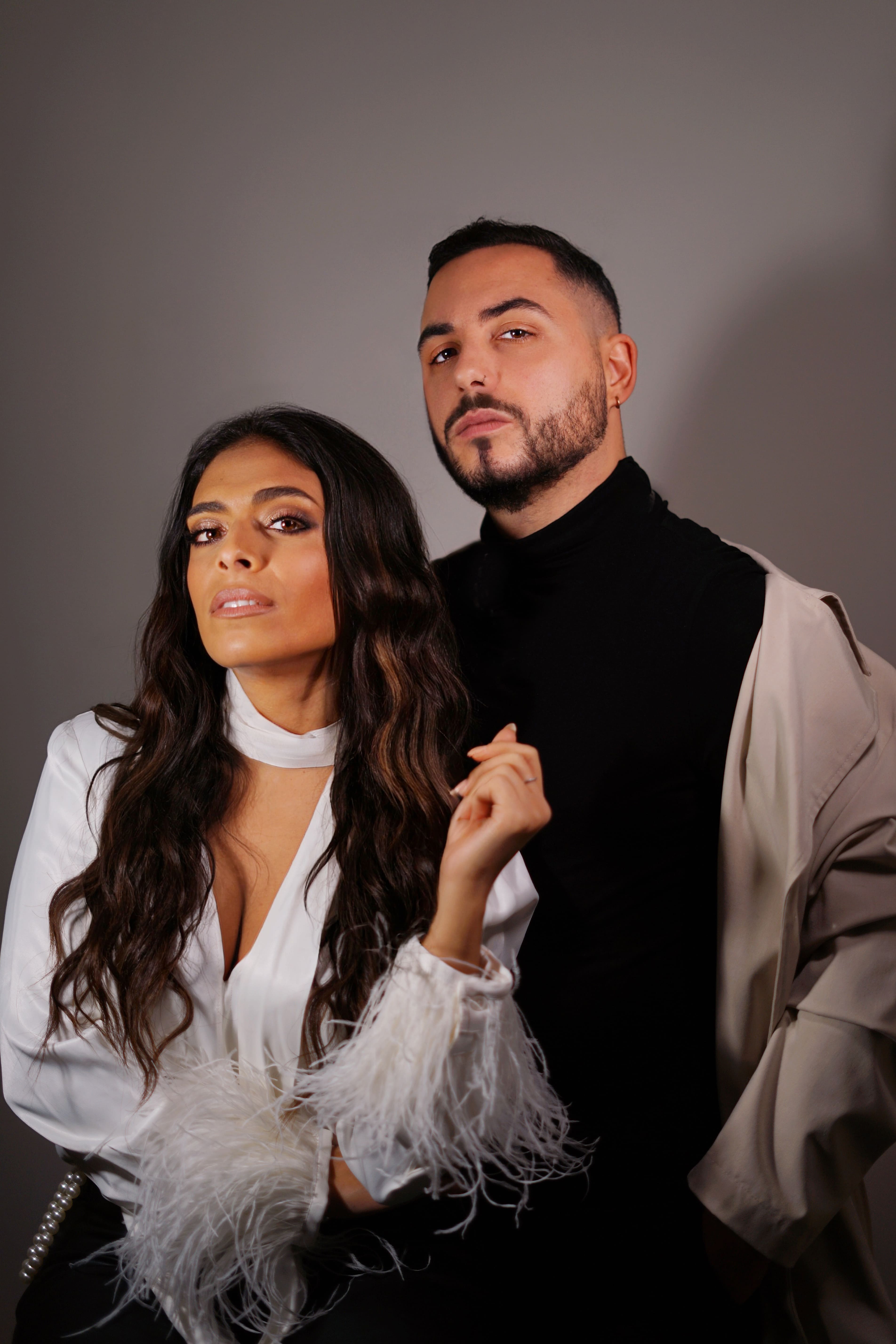 Portrait of a woman with long wavy hair wearing a white blouse with feathered cuffs posing next to a man with short hair, beard, and mustache dressed in a black turtleneck and beige coat.