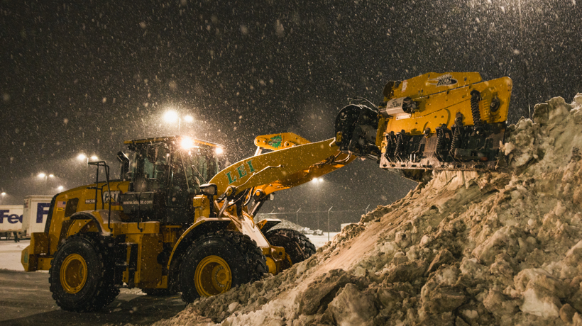 Yellow snowplow loader clearing a large snow pile at night during snowfall.