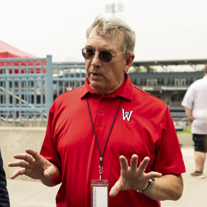 Man wearing sunglasses and red polo shirt speaking outdoors near blue fencing and stadium seating.
