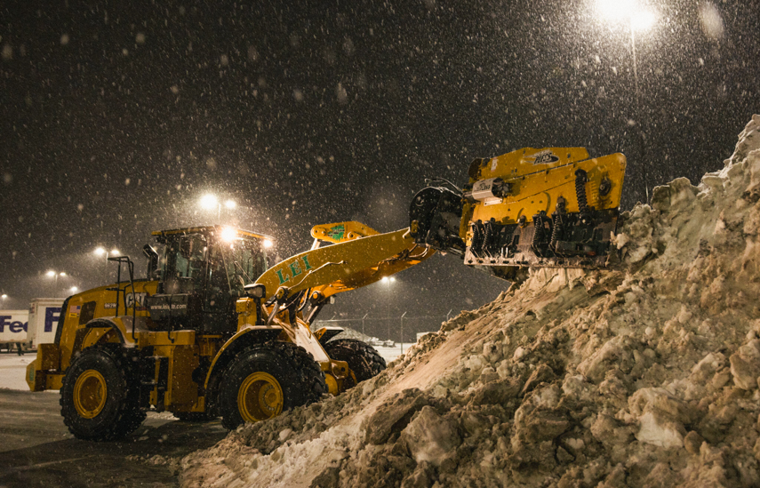 Yellow snowplow clearing a large pile of snow at night with streetlights and falling snow.