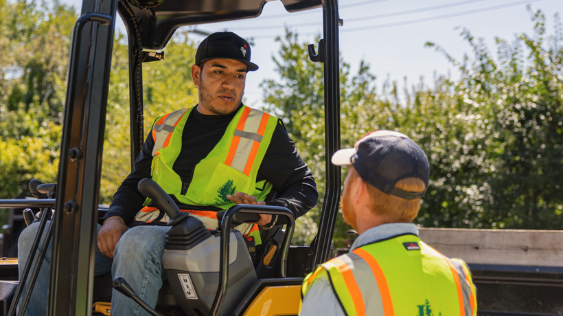 Two construction workers in safety vests talking outdoors near machinery on a sunny day.