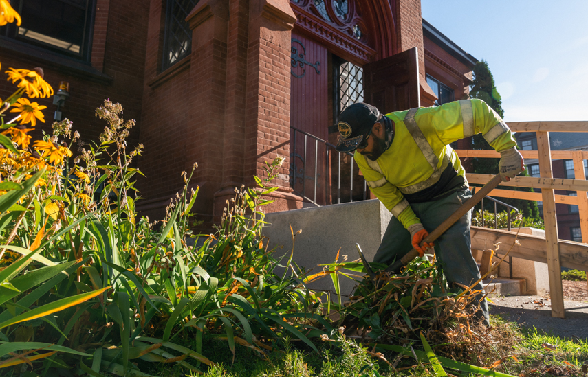 Gardener in yellow safety jacket pulling plants outside a brick building with red doors.