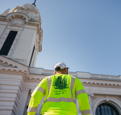 Worker in bright yellow safety jacket stands facing historic building under clear blue sky.
