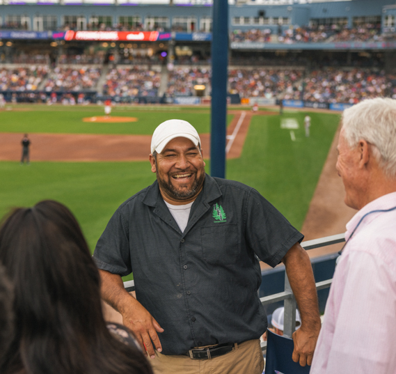 Smiling man in a baseball stadium talking with two people near railing in front of the field.