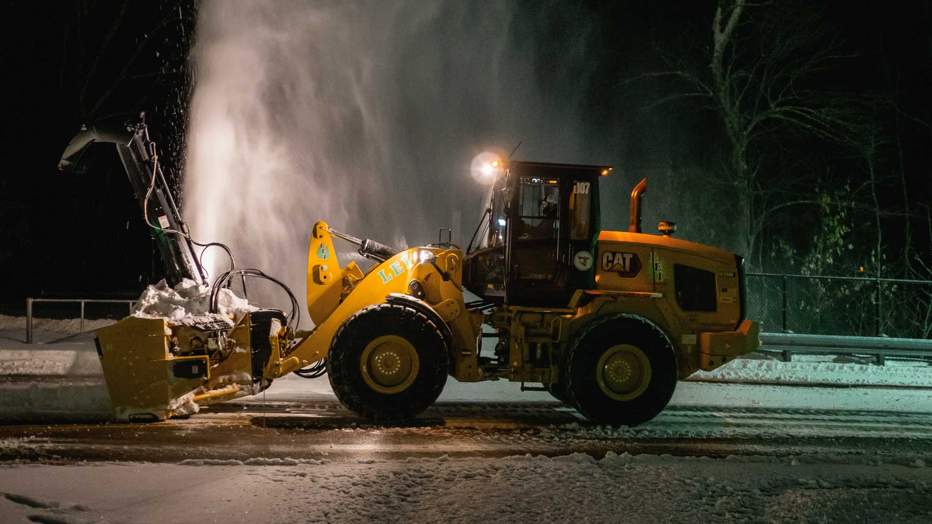 Yellow CAT snowplow clearing snow on a road at night with snow spraying upward.