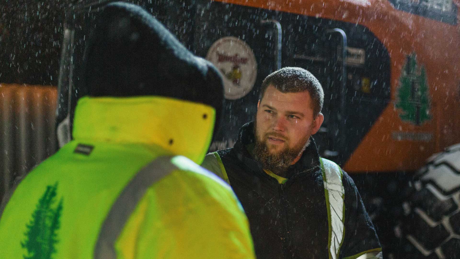 Two workers in rainwear talk outside at night with snow falling around them.