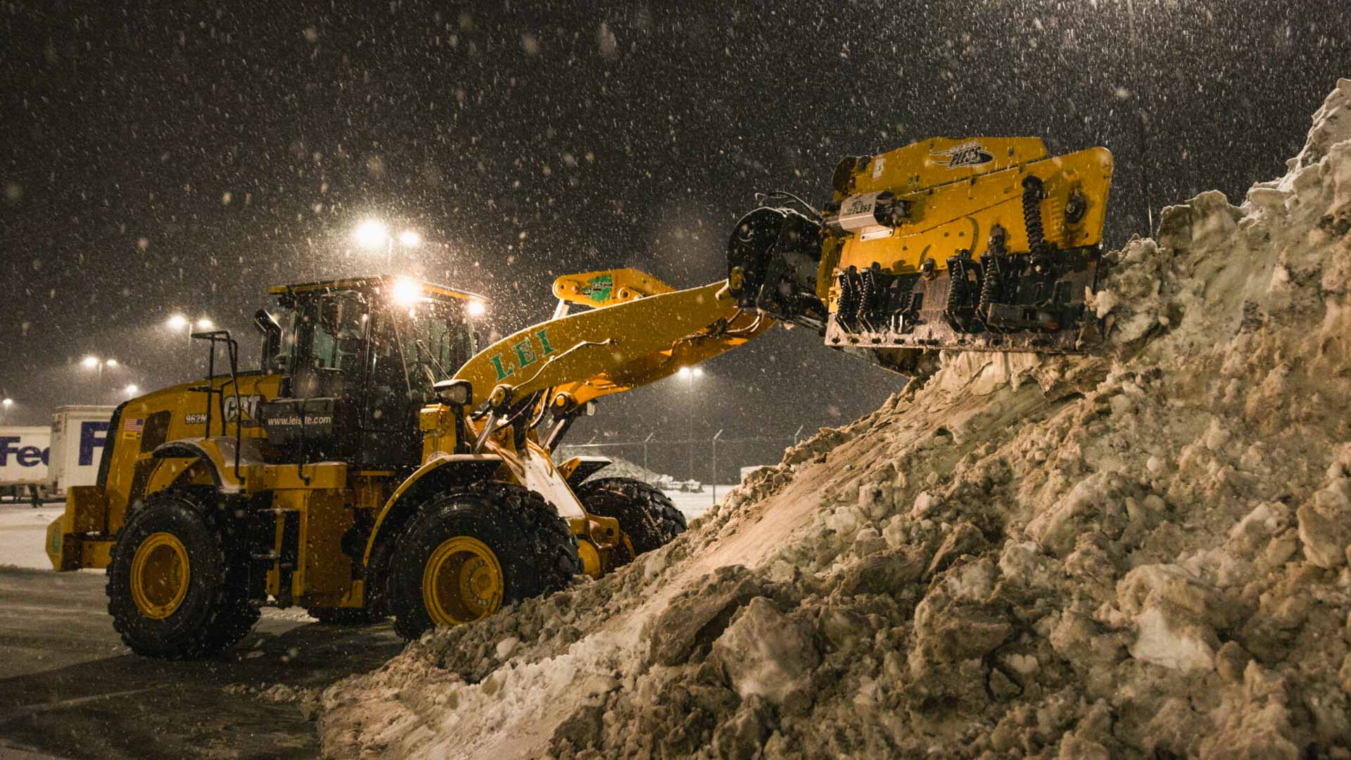 Yellow front loader clearing a large dirty snow pile at night in a snowstorm.
