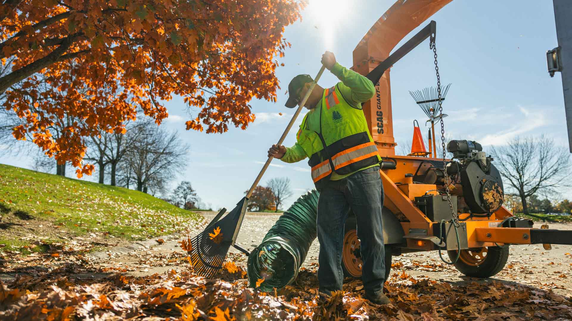 Worker in fluorescent vest raking autumn leaves into a leaf vacuum machine outdoors.