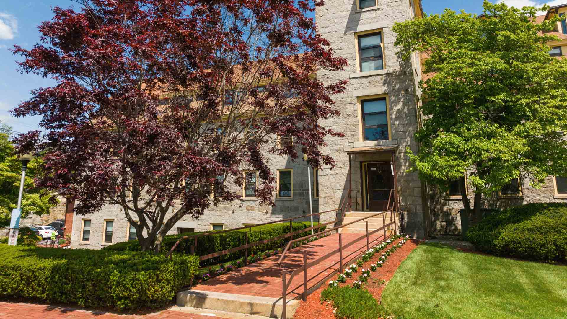 Stone building with ramp entrance, red-leaf tree on left, green tree on right, and manicured lawn.