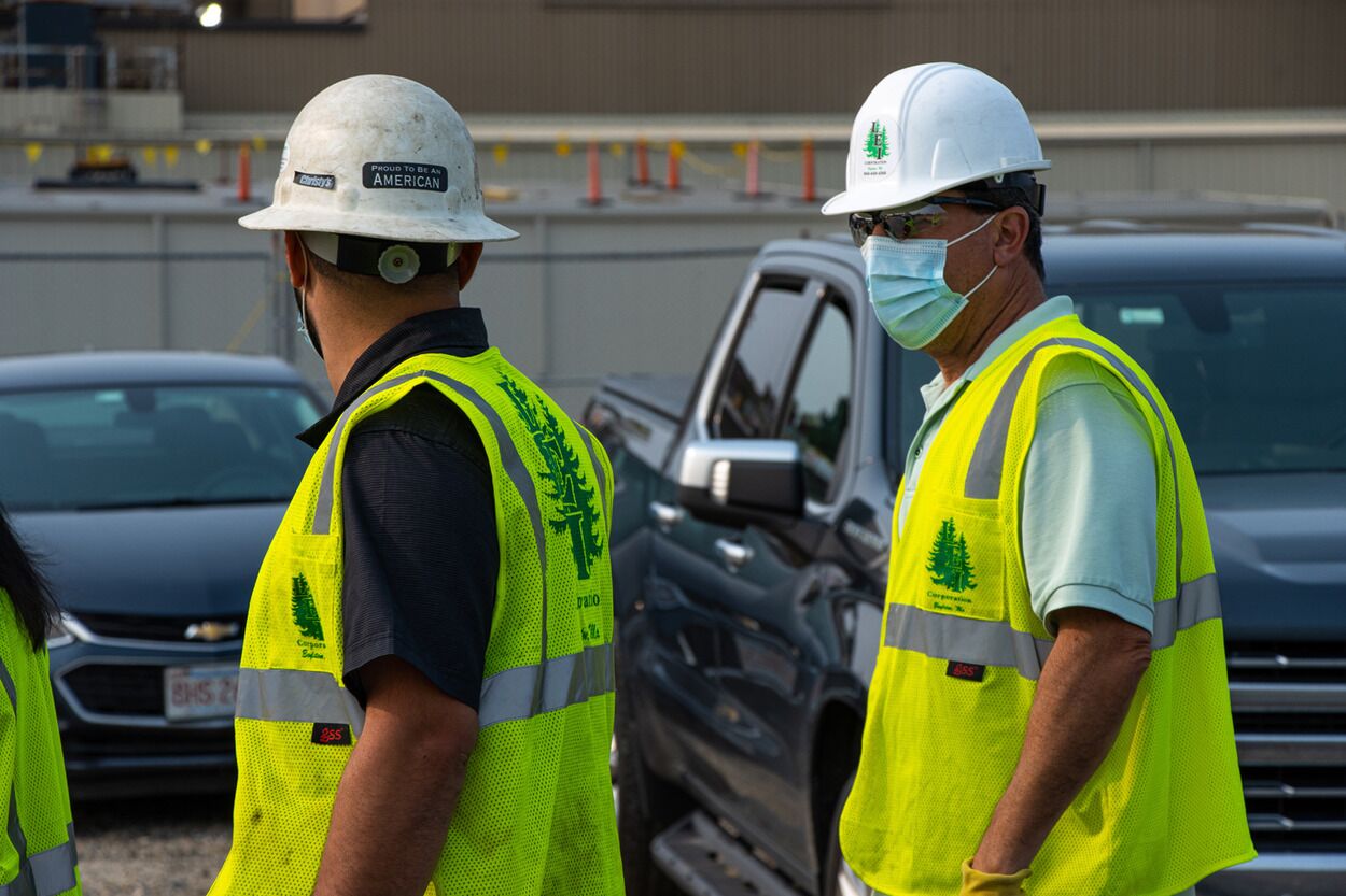 Two construction workers in yellow vests and white helmets, one wearing a face mask, near parked vehicles.