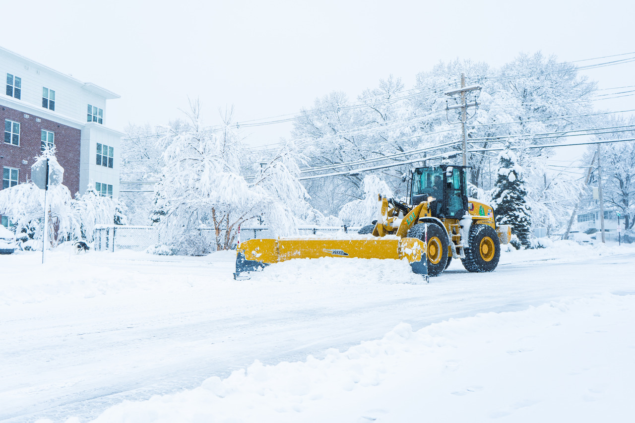Yellow snowplow clearing snow on a street lined with snow-covered trees and buildings.