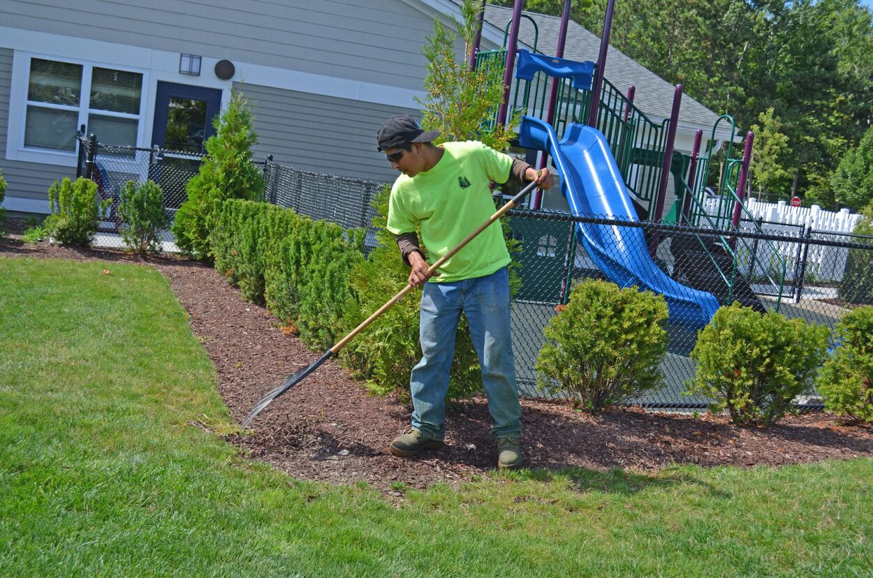 Man wearing a neon green shirt rakes mulch around green shrubs near a playground slide.