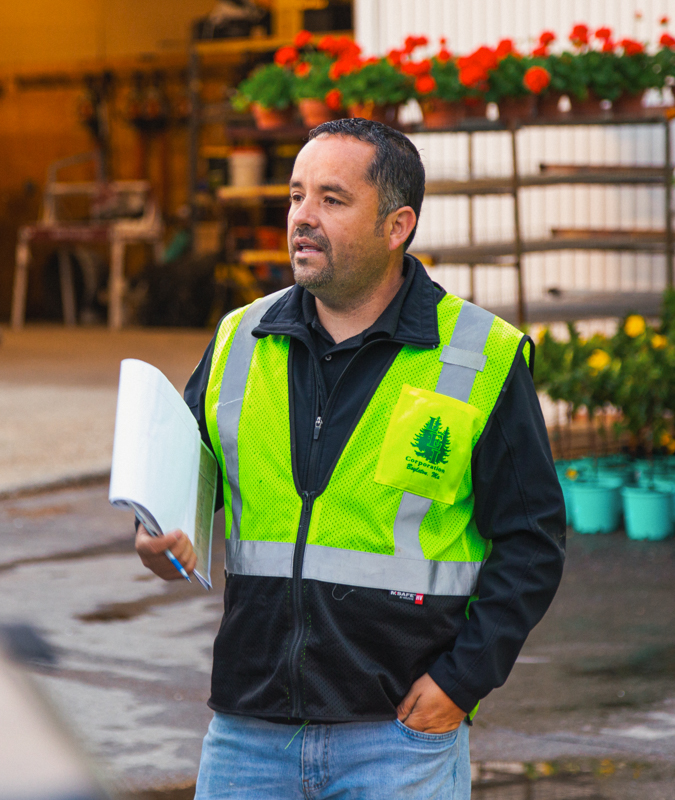 Man in a neon yellow safety vest holding papers stands near flower pots and shelves in a garden center.