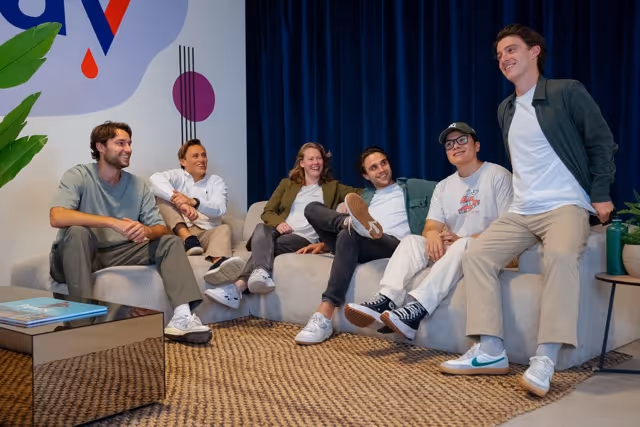 Six casually dressed people sitting and smiling together on a beige couch and chair in a modern room with a blue curtain and abstract wall art.