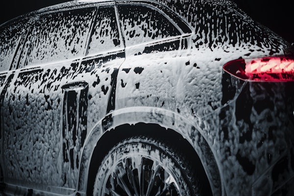 Close-up of a black car covered in thick white foam during a wash, focusing on the rear side and wheel.