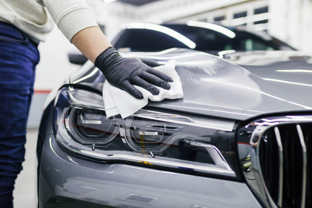 Person wearing black gloves cleaning the hood and headlight of a silver car with a white cloth in a garage.
