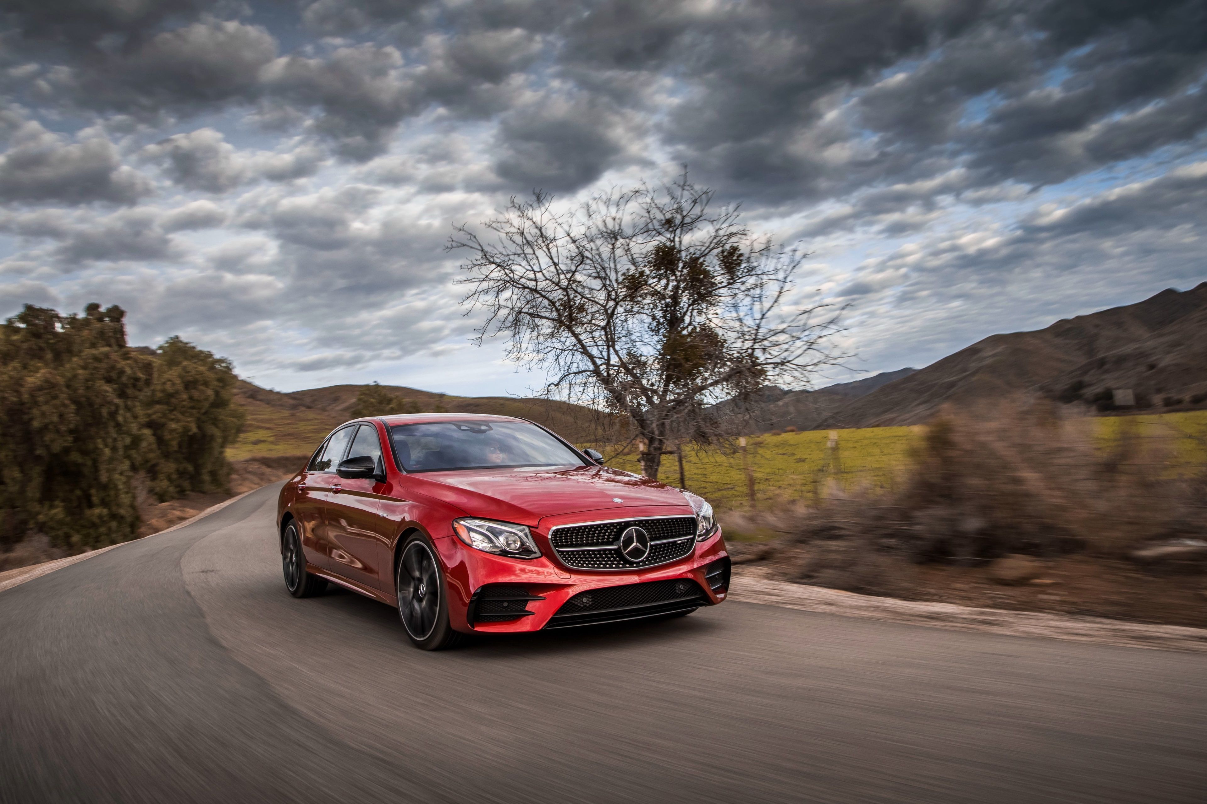 Red Mercedes-Benz sedan driving on a winding road with cloudy sky and mountainous landscape.