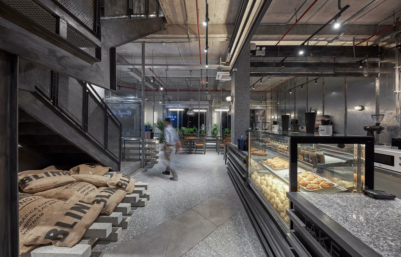 Modern coffee shop interior with glass display case of pastries, sacks of coffee beans near a staircase, and a blurred person walking inside.