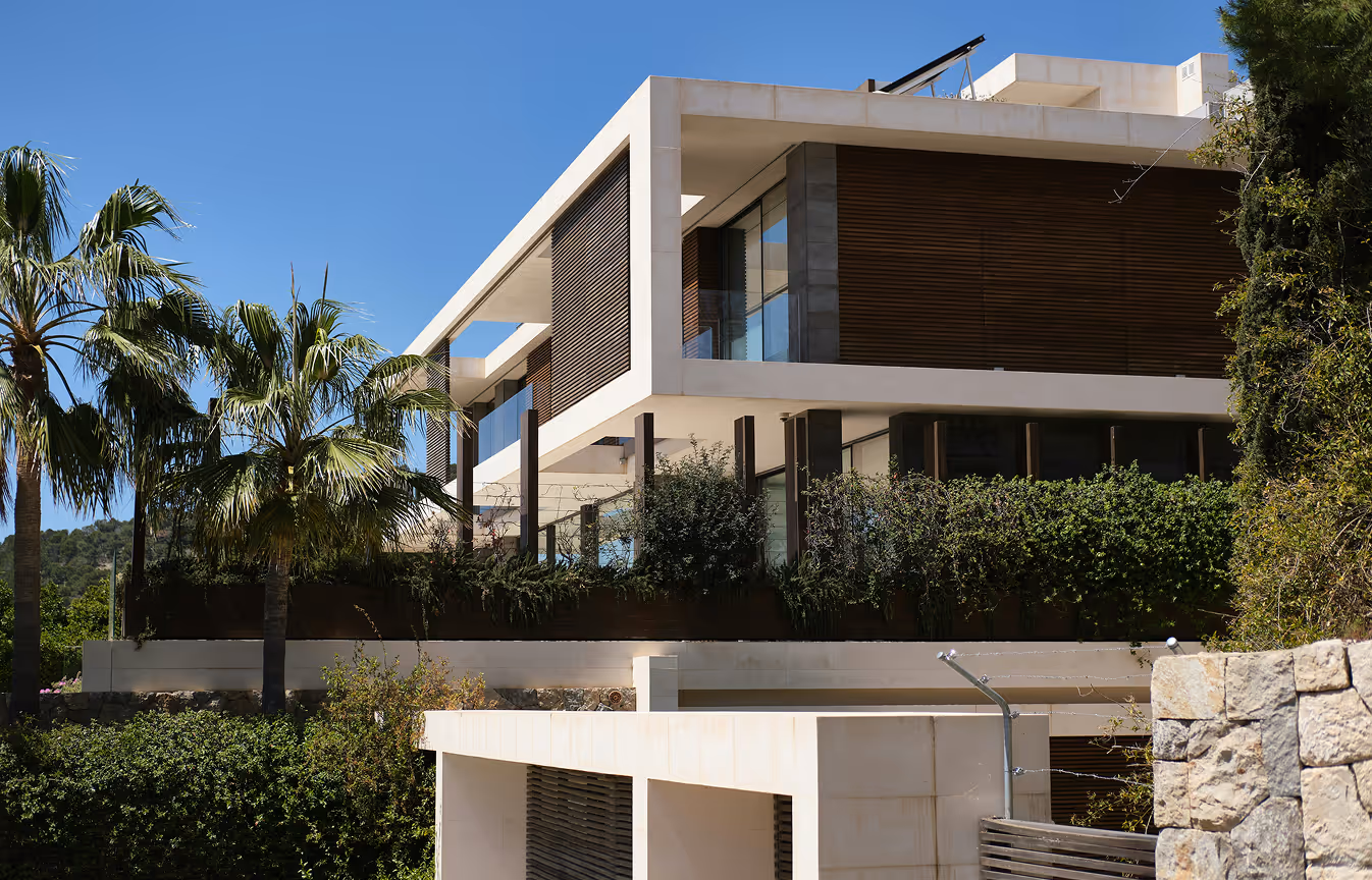Modern multi-story house with large windows, white and dark panel exterior, surrounded by green trees and bushes under a clear blue sky.