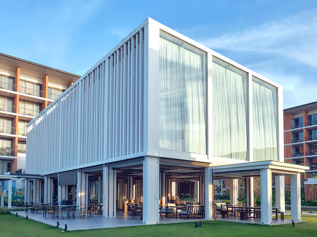 Modern white rectangular building with large glass windows and outdoor seating under an open ground level, set against a blue sky.