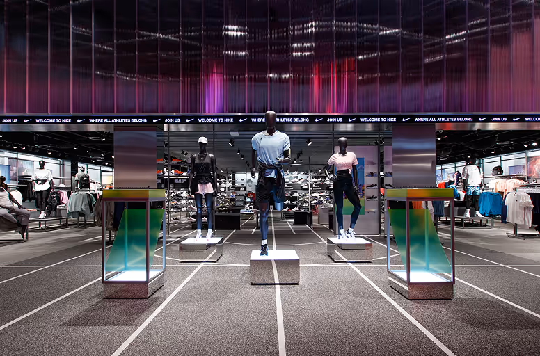 Nike store display with three mannequins dressed in athletic wear on pedestals, surrounded by apparel racks and a large digital screen overhead.