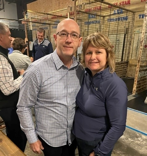 Man in checkered shirt and glasses standing next to woman in navy jacket, both smiling indoors.