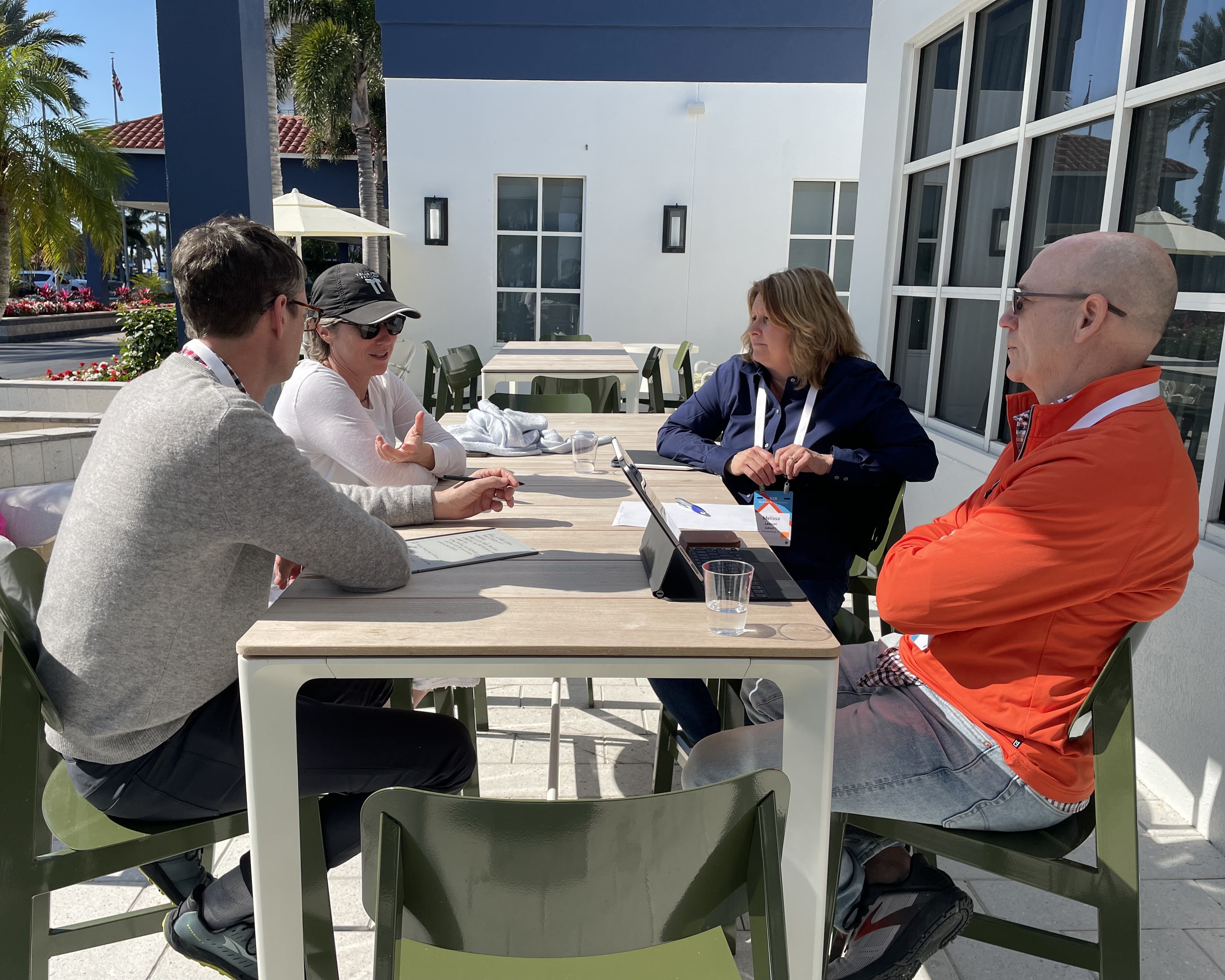 Four adults seated outdoors at a table in discussion, with laptops and papers on the table.