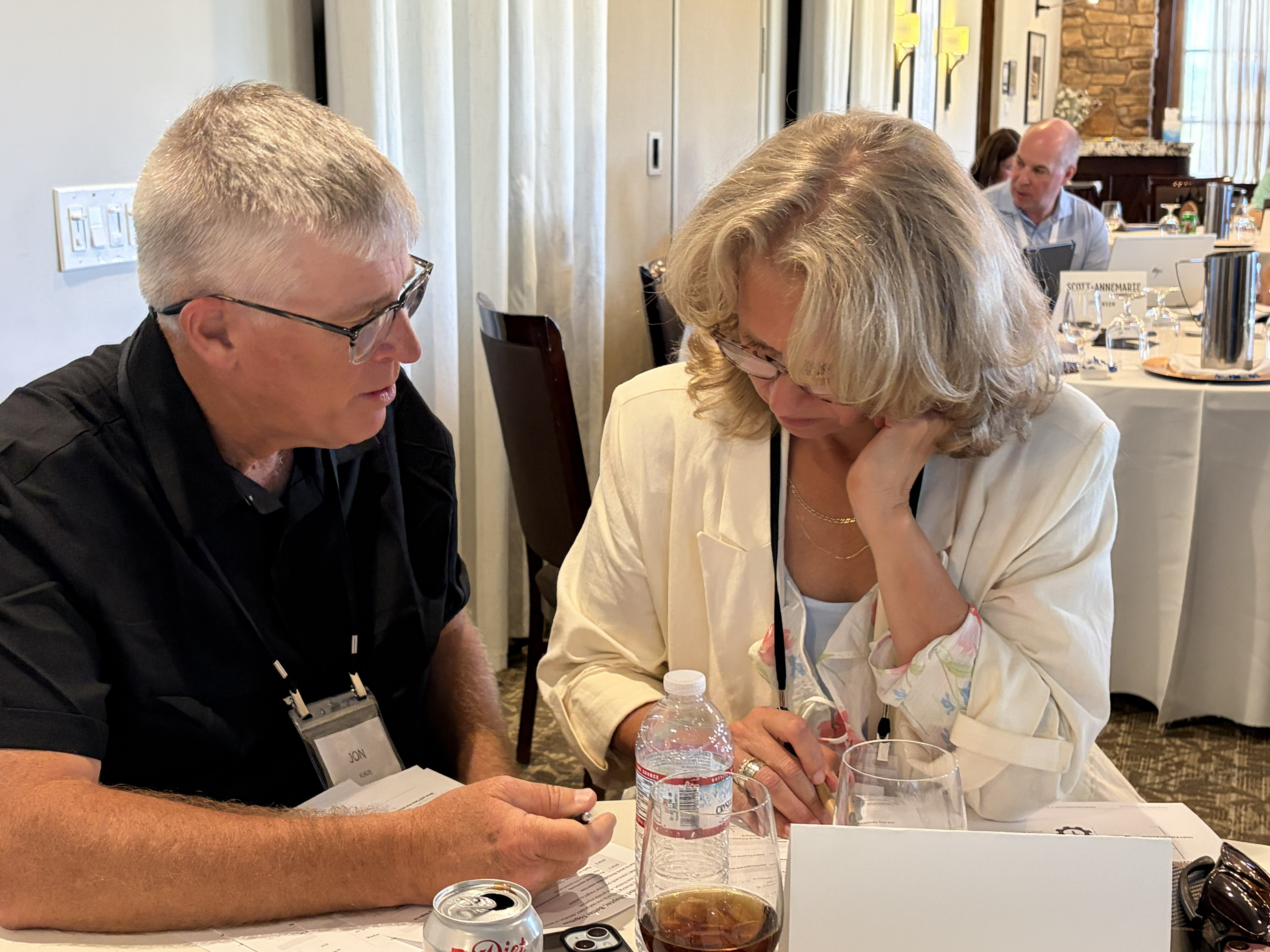Man and woman sitting at table, engaged in discussion with papers and drinks in front of them.