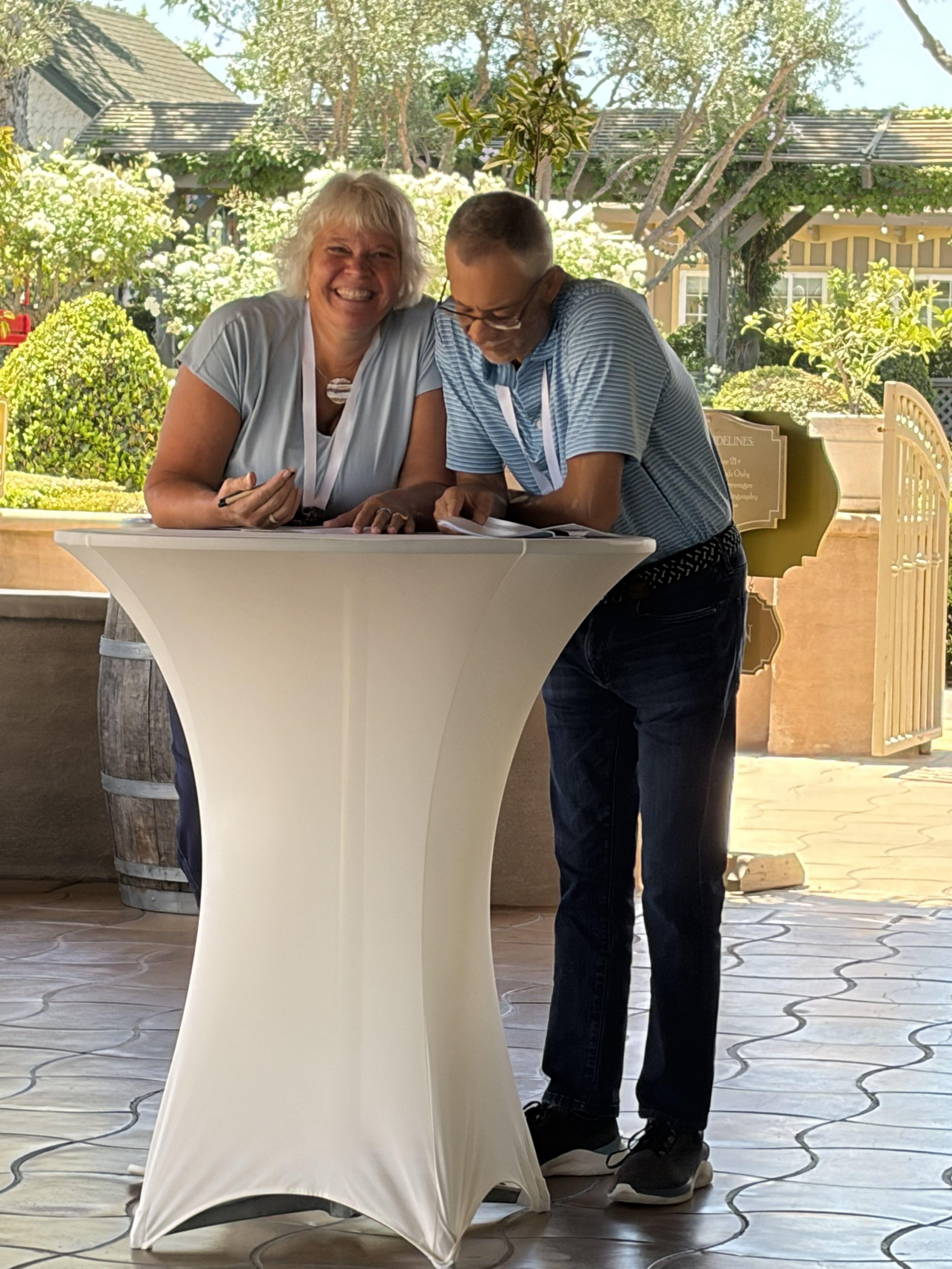 Smiling couple standing at a tall white table outdoors, writing or reading papers together.
