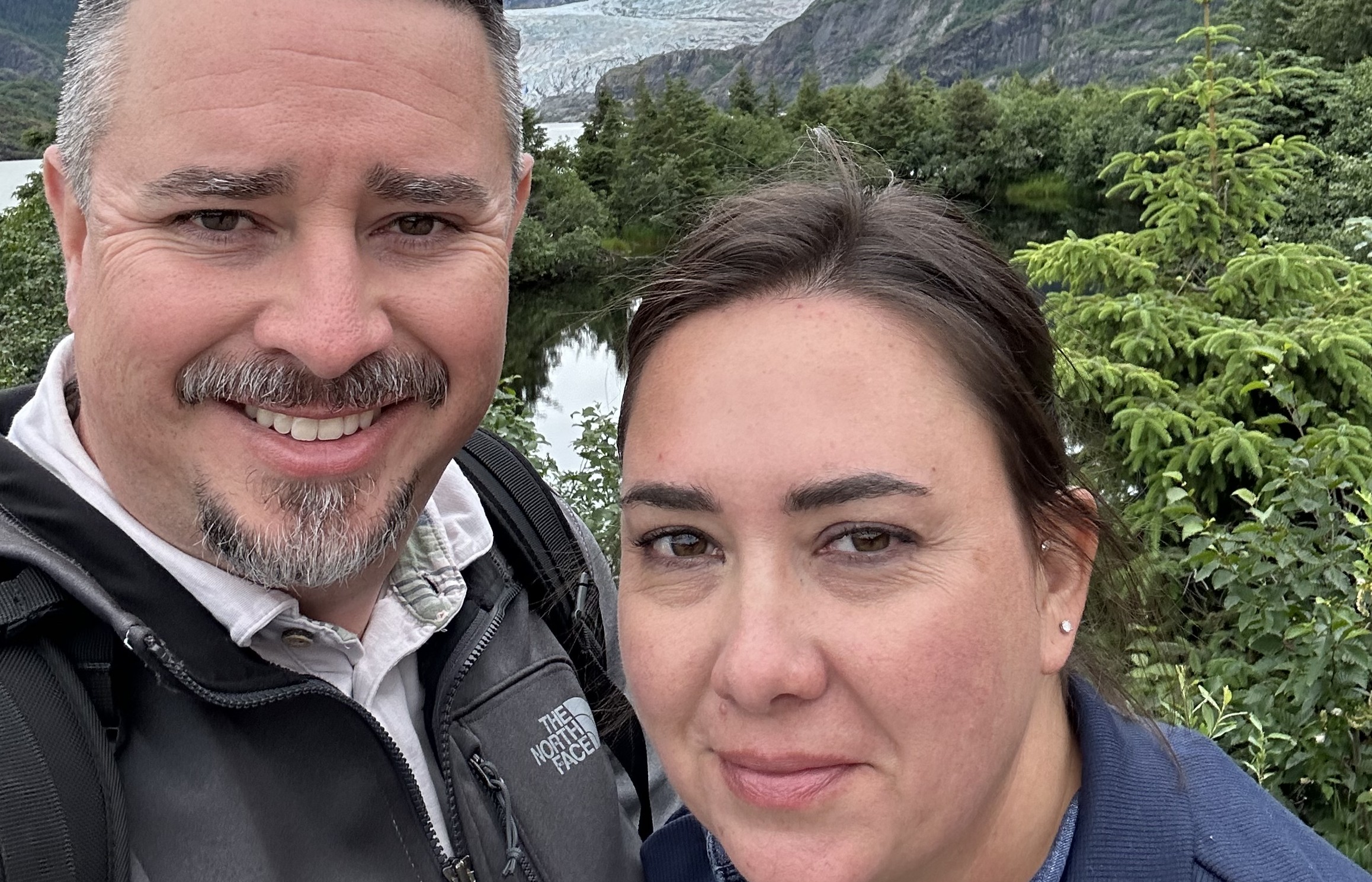 Close-up selfie of a smiling man and woman outdoors with trees and a mountain in the background.