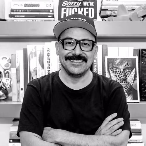 Mann mit Schnurrbart, Brille und Baseballcap steht lächelnd vor einem Bücherregal mit bunten Büchern.