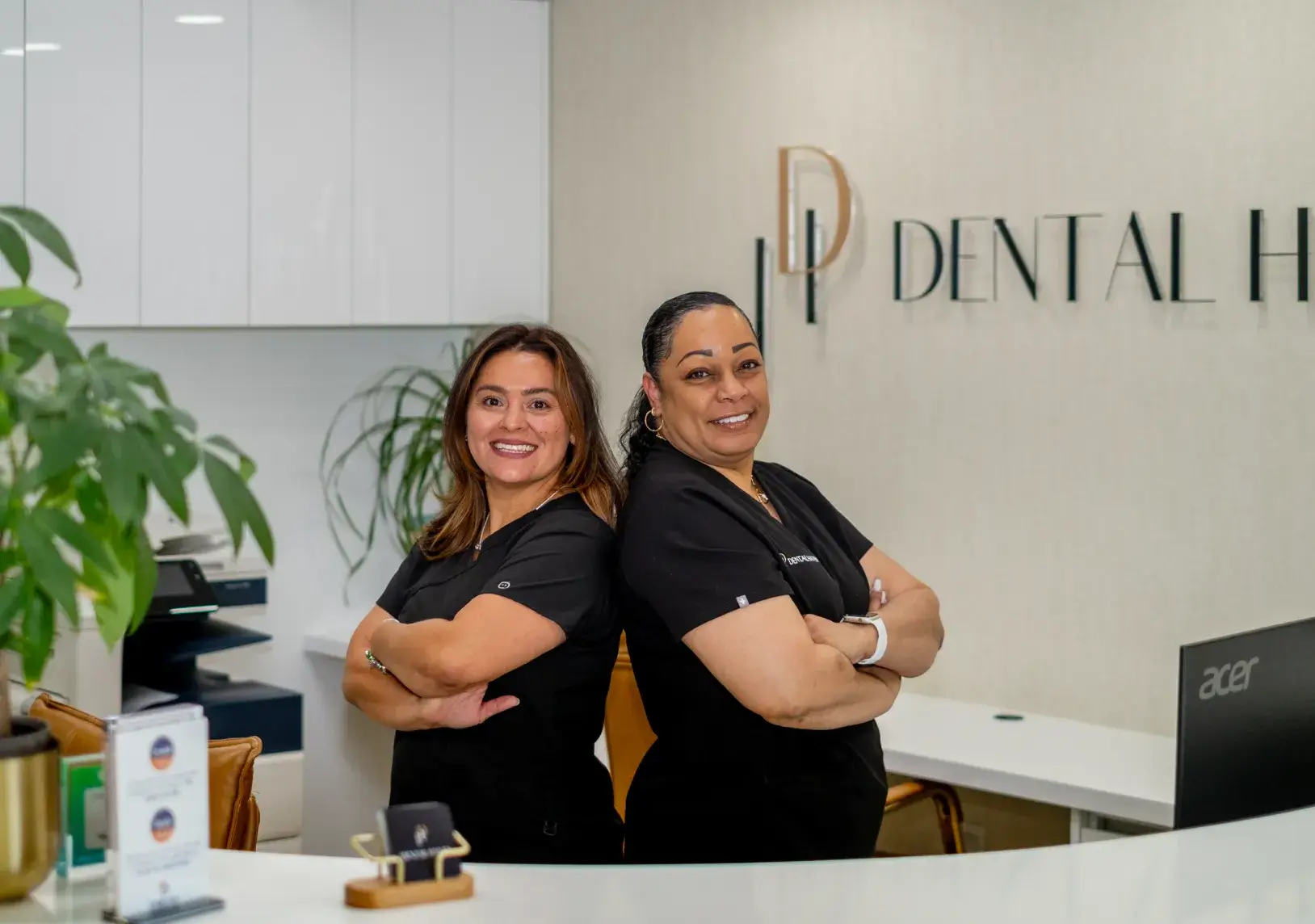 Two smiling dental professionals stand back-to-back in a dental office, wearing black uniforms.