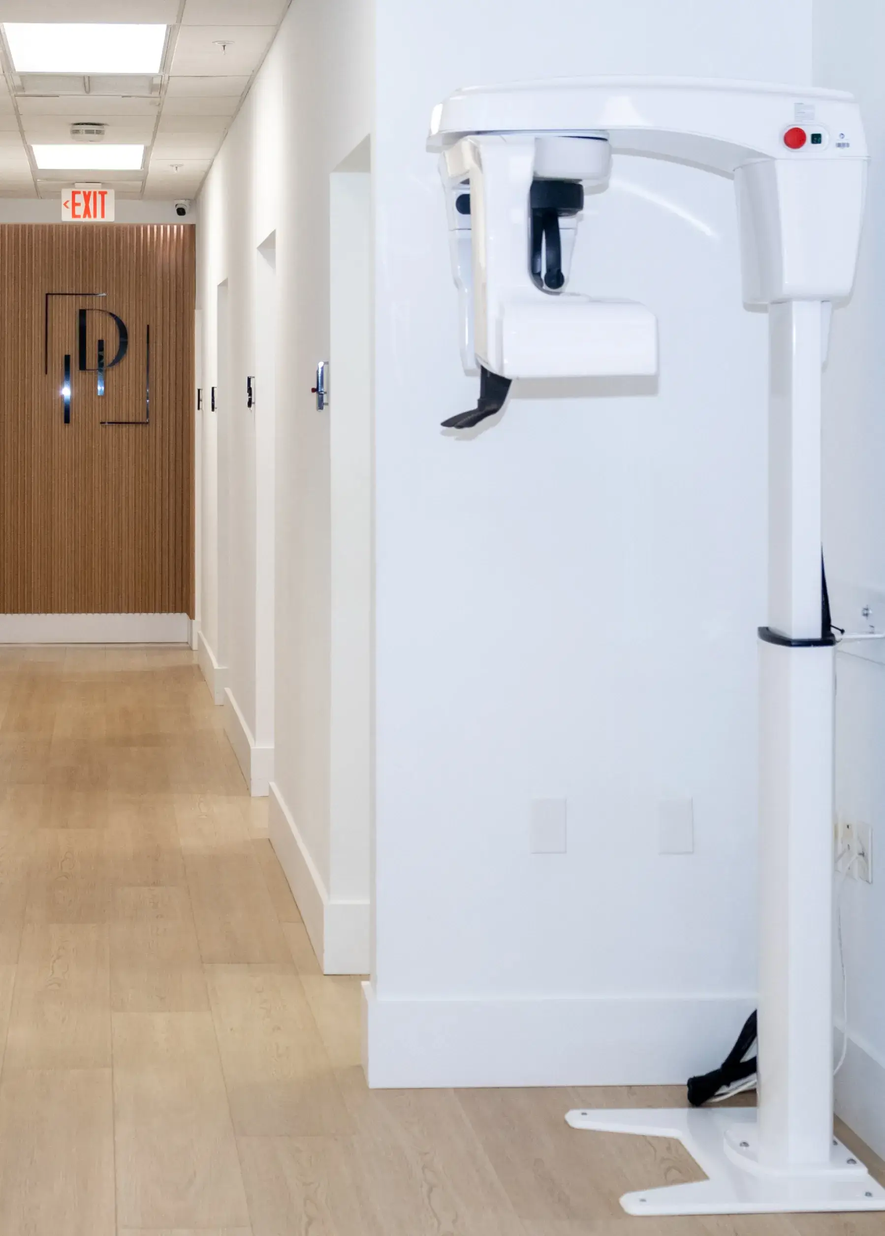 A dental X-ray machine stands in a hallway near white walls and wooden flooring.
