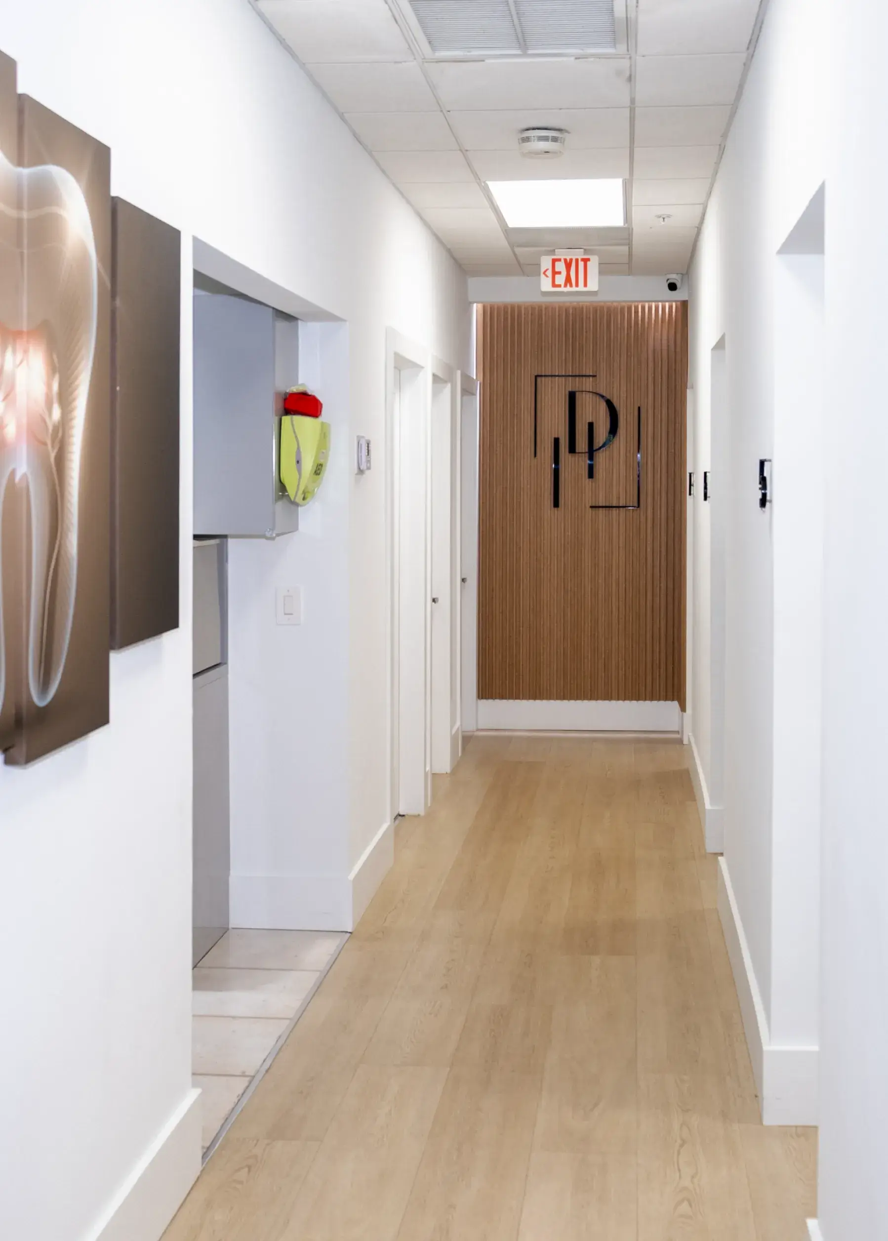 A hallway with medical-themed artwork on the walls, leading to a wooden door with an exit sign above.