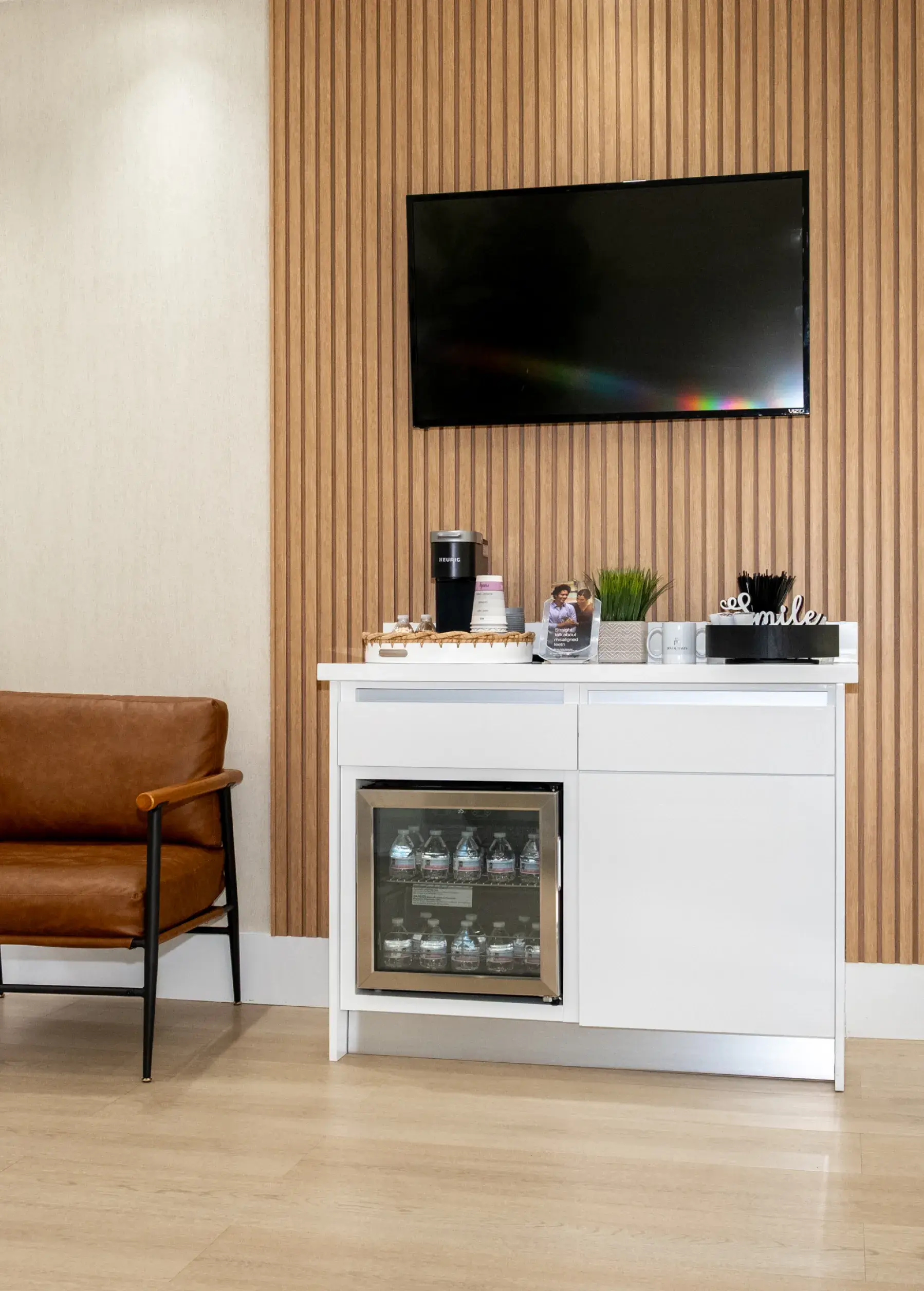 A white cabinet with a mini-fridge and coffee machine is next to a brown chair.