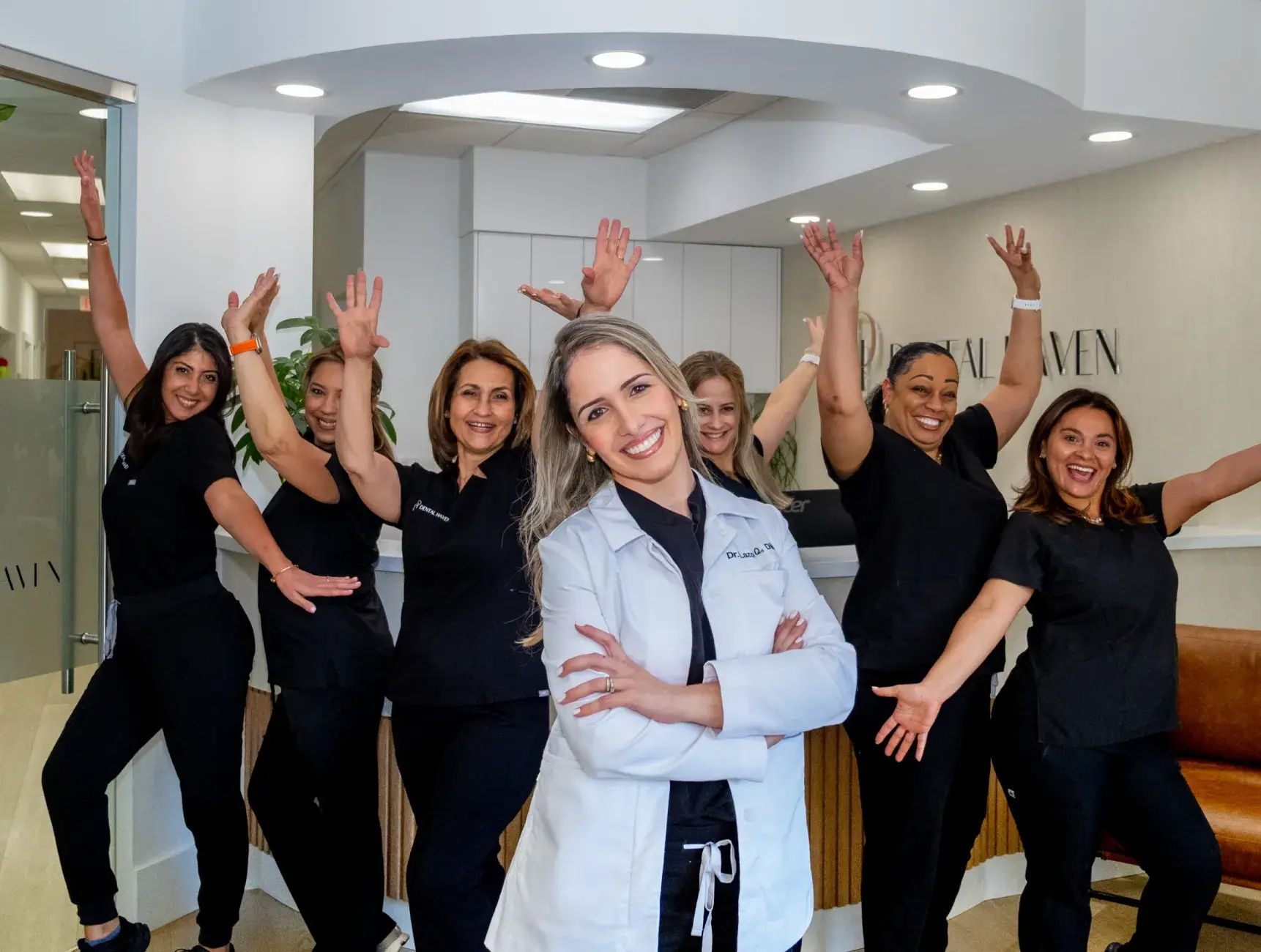 A smiling woman in a white coat stands in front of five happy women in black uniforms.