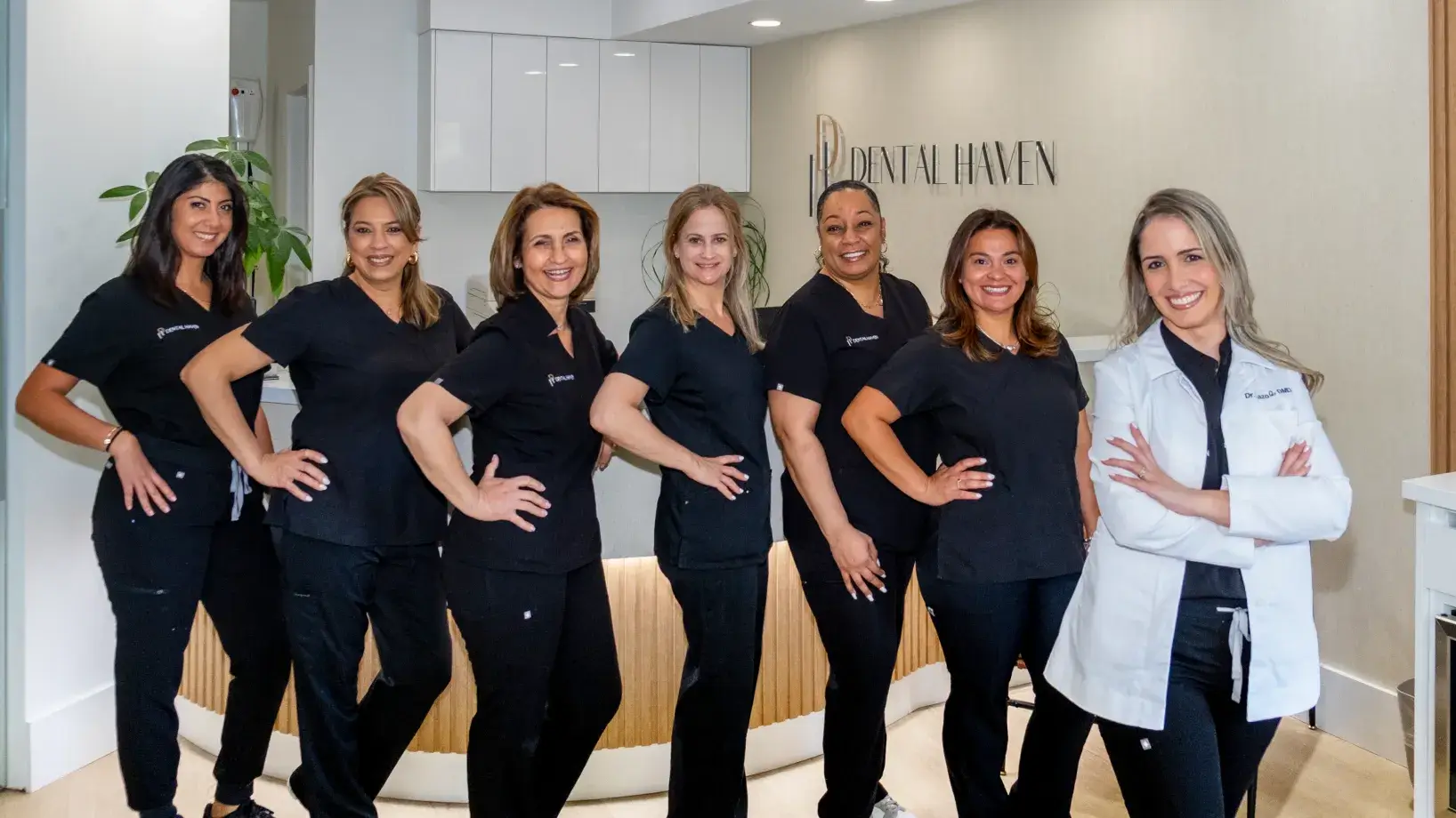 Seven women in black scrubs and a smiling woman in a white coat stand together at a dental office.
