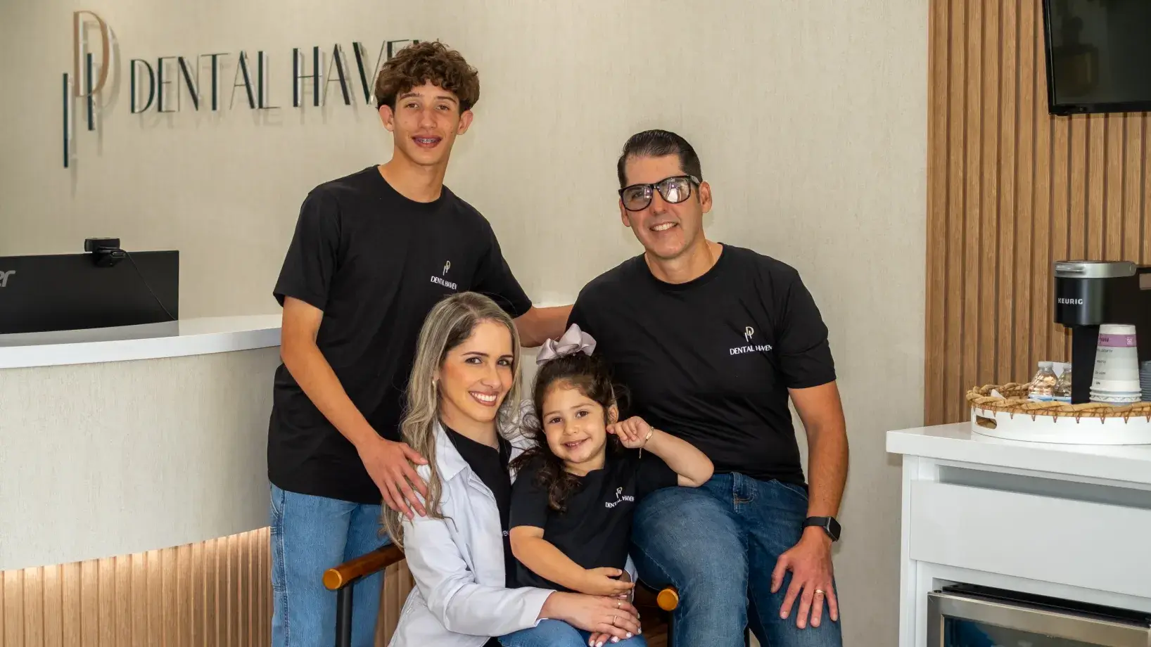 A family of four stands together in a dental office, all wearing black shirts and smiling.