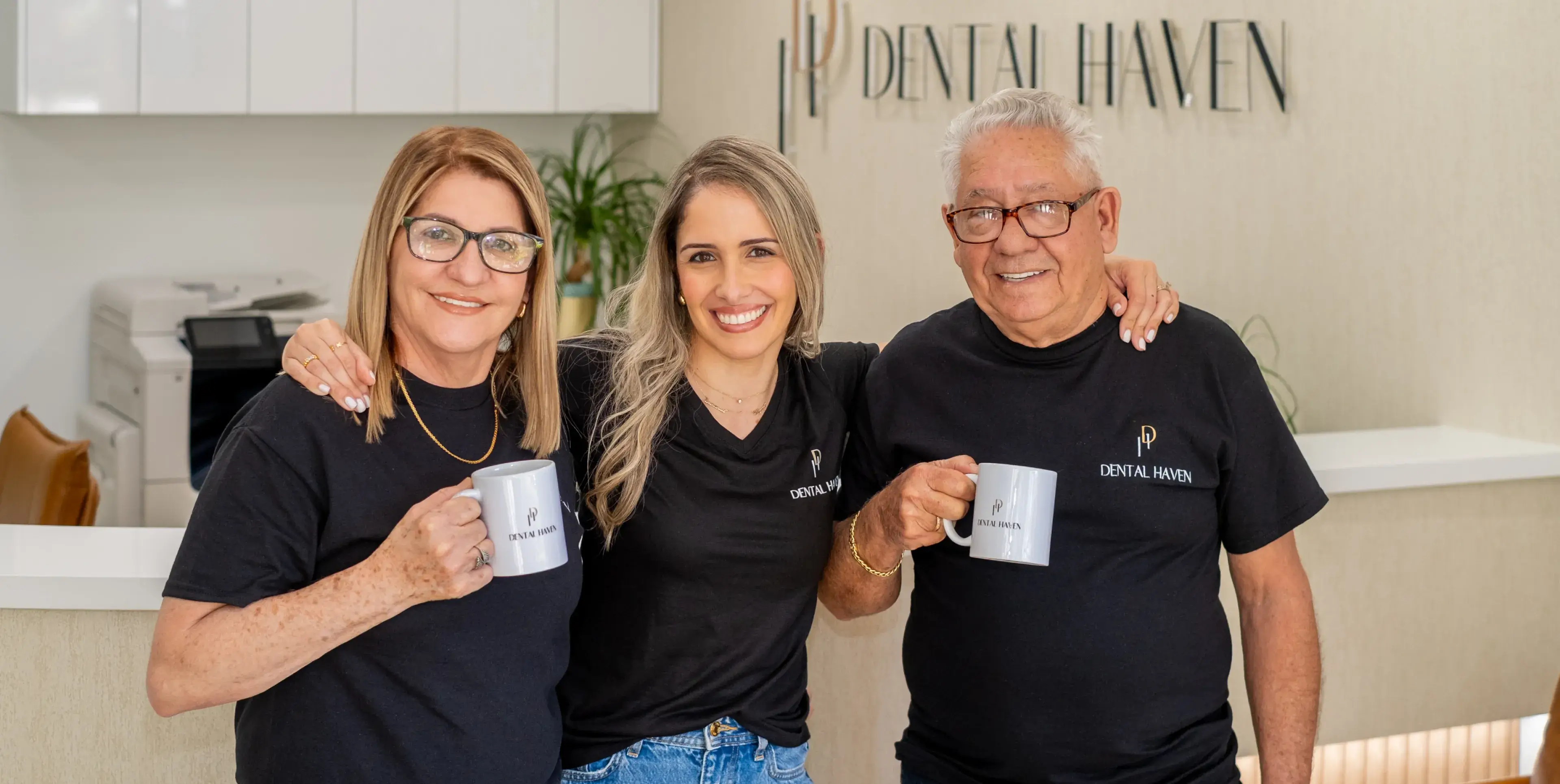 Three people in matching shirts stand together, smiling, and holding coffee mugs inside a dental office.