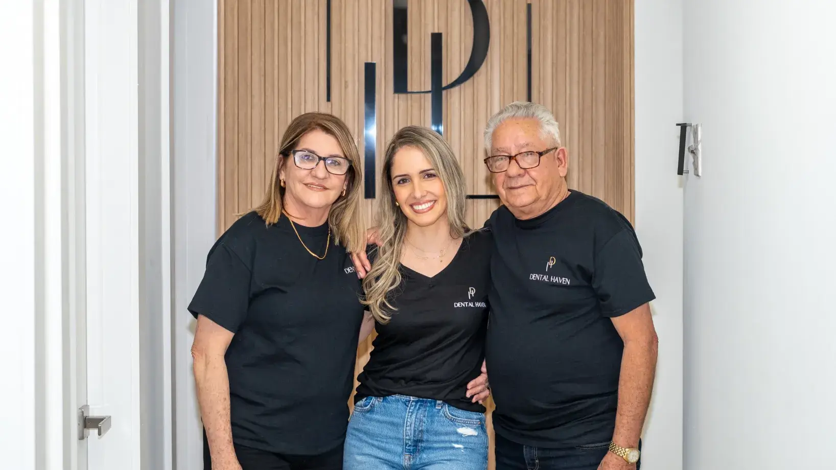 Three people in black shirts posing together and smiling in front of a wooden panel.