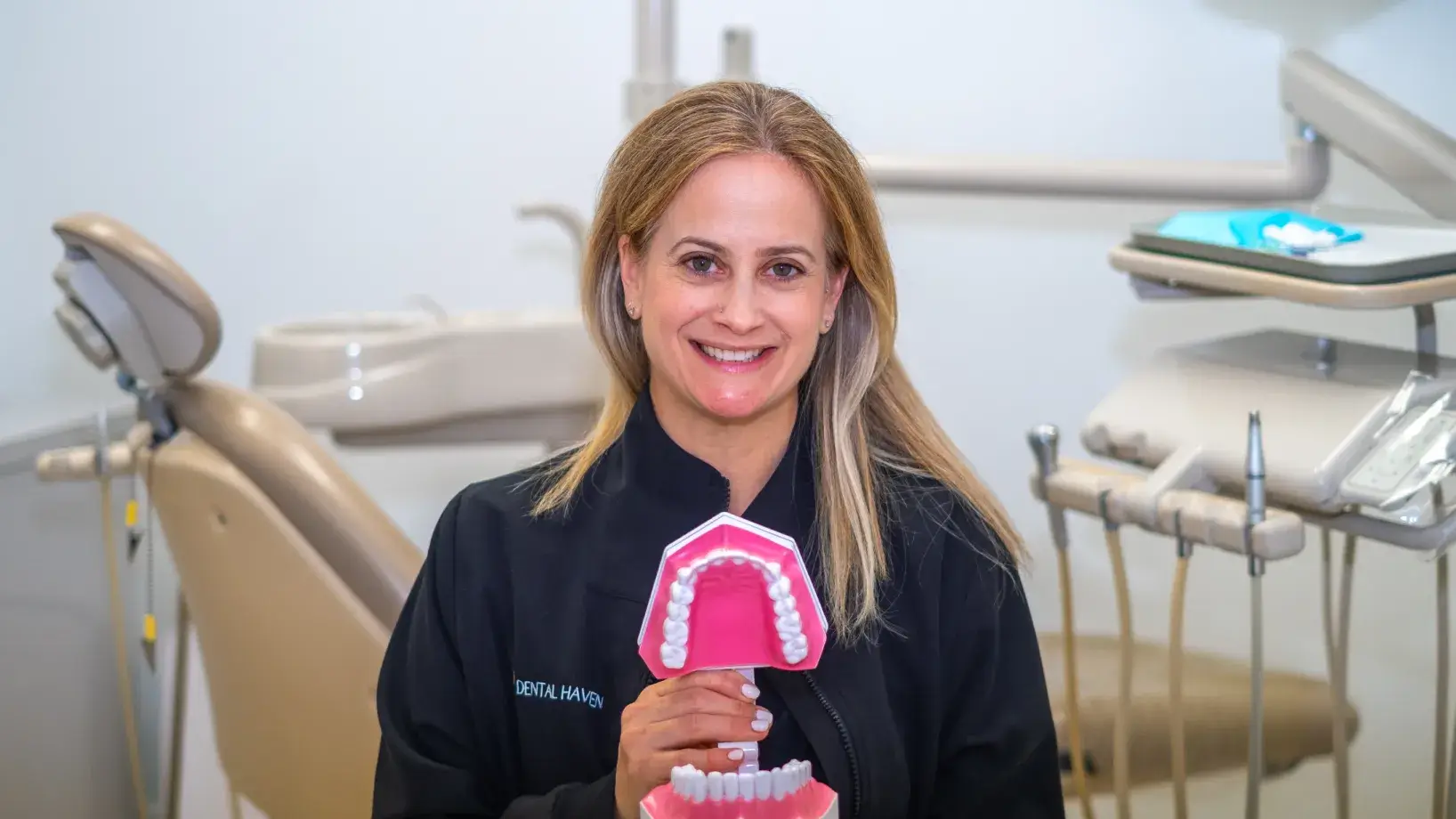 A dentist holding a dental model in an examination room, smiling at the camera.