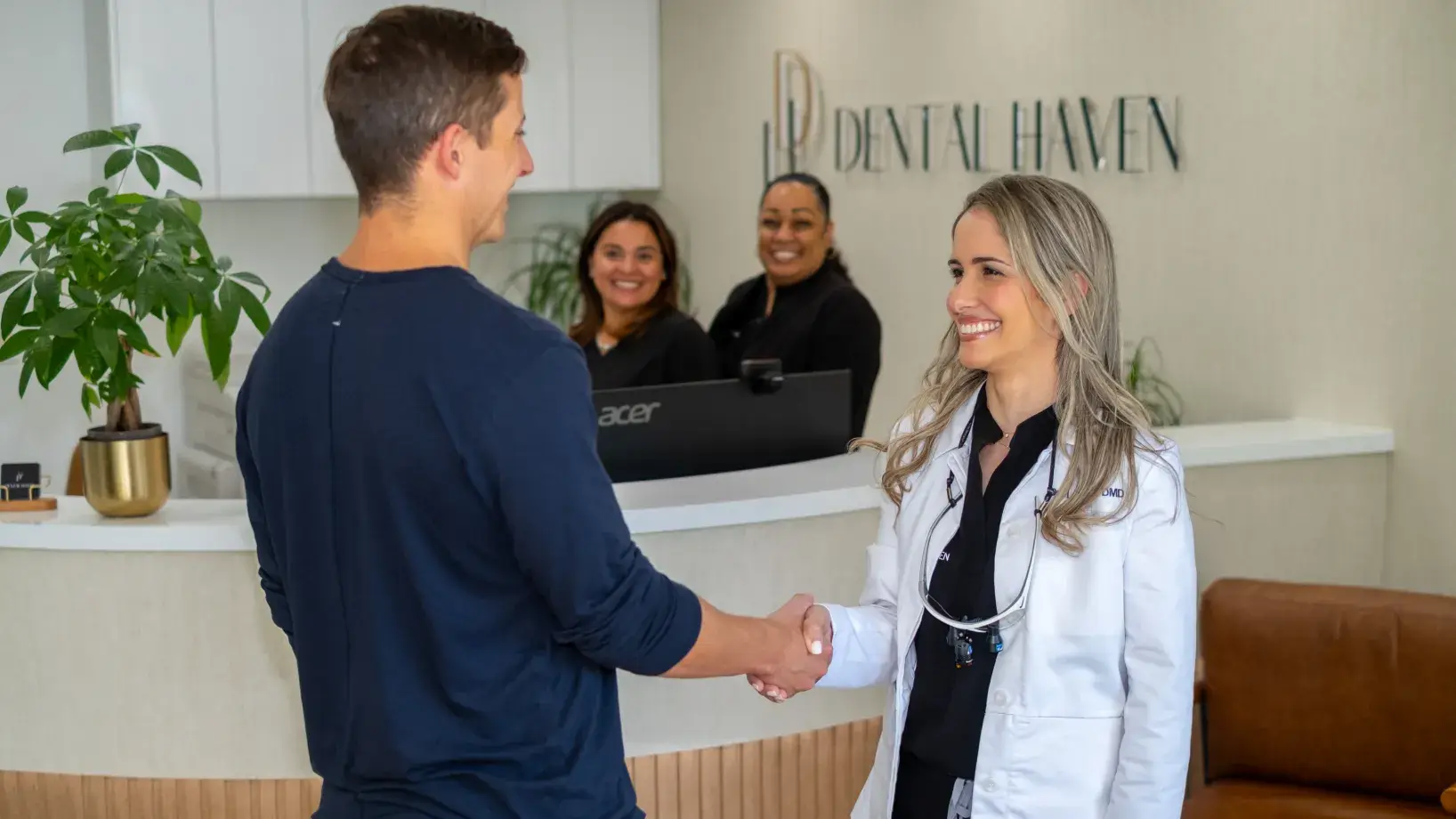 A dentist in a white coat shakes hands with a patient at a dental office reception.