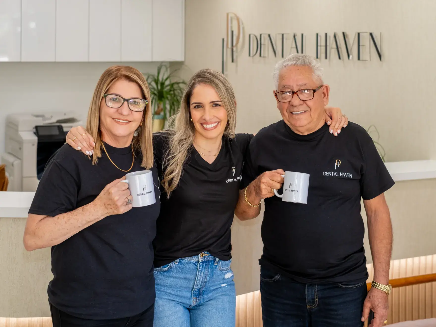 Three smiling people holding mugs, standing in front of a "Dental Haven" reception area.